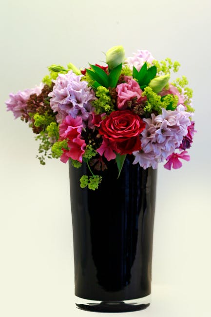 A vibrant floral arrangement featuring a variety of fresh flowers in a tall, sleek black vase. The bouquet includes prominent red roses, soft pink hydrangeas, pale green lisianthus buds, and small spr