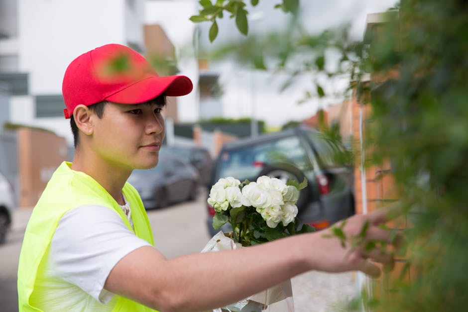 A young male flower delivery courier, wearing a red cap and high-visibility yellow vest over a white t-shirt, is seen placing a bouquet of fresh white hydrangeas and roses against a green leafy wall o