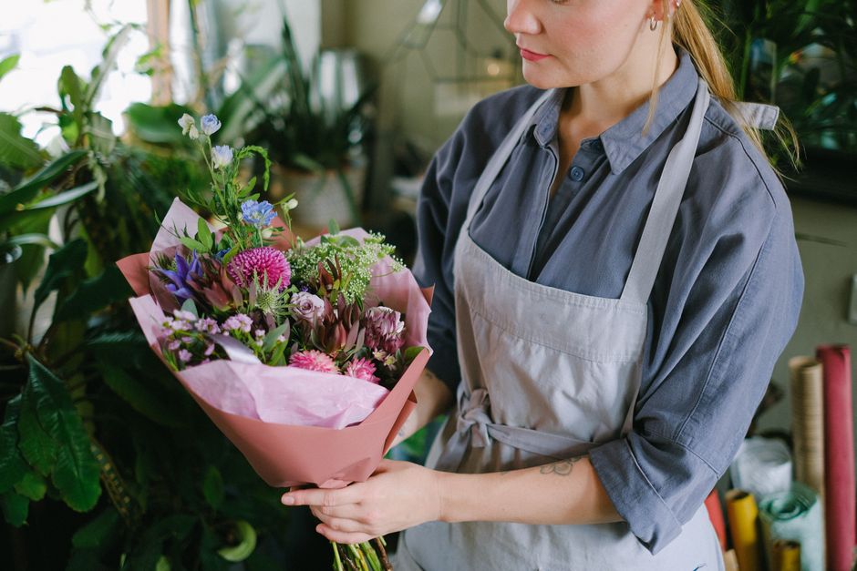 A woman in a grey shirt and light grey apron, standing in a floral workshop, holding a vibrant bouquet of fresh flowers wrapped in pink tissue paper. The bouquet features a mix of pink and purple flow