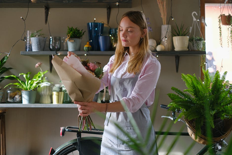 A young woman with long, light brown hair wearing a white shirt and light gray apron is holding a large bouquet of mixed flowers wrapped in brown paper. The bouquet includes pink roses, white peonies,