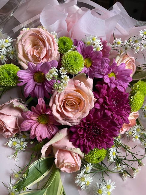 A vibrant floral bouquet featuring pale pink roses, purple chrysanthemums, bright green button mums, and small white daisies with yellow centers, all arranged with green foliage and wrapped in delicat