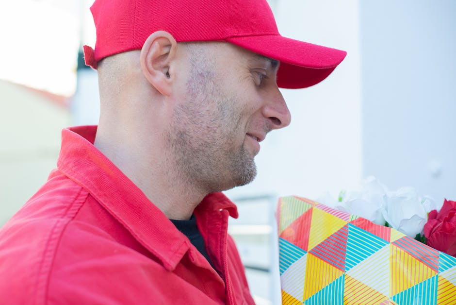 A close-up profile view of a man wearing a red cap and matching red jacket, holding a colorful gift bag with a geometric pattern in red, yellow, blue, and white, filled with white and pink flowers, po