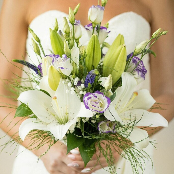 A moment of serene celebration is depicted as a bride stands in Bexleyheath, cradling an exquisitely composed bouquet, a cascade of white and purple set against the classic silhouette of her strapless wedding dress. Several large, radiant white Oriental lilies command attention, their petals velvety and luminous, delicately brushed with subtle spots and boasting golden-orange stamens. Nestled between the lilies are fresh lisianthus blossoms, crisp and airy, with hints of lavender tracing the edges, their presence alongside unopened buds promising a story unfolding. Slender clusters of deep violet-blue Veronica flowers add a vibrant accent, echoing the colorful spirit found at local gatherings in Bexleyheath's parks. Delicate baby's breath floats above the arrangement, lending an ethereal softness, while generous emerald foliage - broad lily leaves and whispery asparagus fern - creates an embrace of green around the florals. At the base, the bride's hands adorned with light, neutral polish gently clasp the stems, their grace mirrored by the bouquet itself. The lush tones, textural contrasts, and strategic design reflect the work of seasoned local florists, offering this bouquet as a symbol of romance and refined taste for celebrations across Bexleyheath, whether marking moments near Bostall Woods or cherished weddings at local venues.